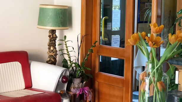 Un suelo a cuadros, un sofá rojo y blanco, una lámpara, una puerta y un jarrón con flores.