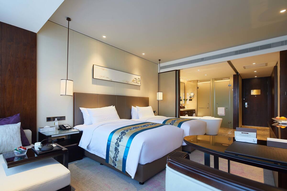 A hotel room with two beds, a sitting area with a table, a mirror, and a bathroom visible through an open door.