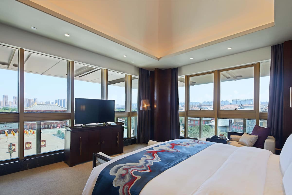 A hotel room with a large bed, a TV on a wooden cabinet, and a view of a cityscape through large windows.