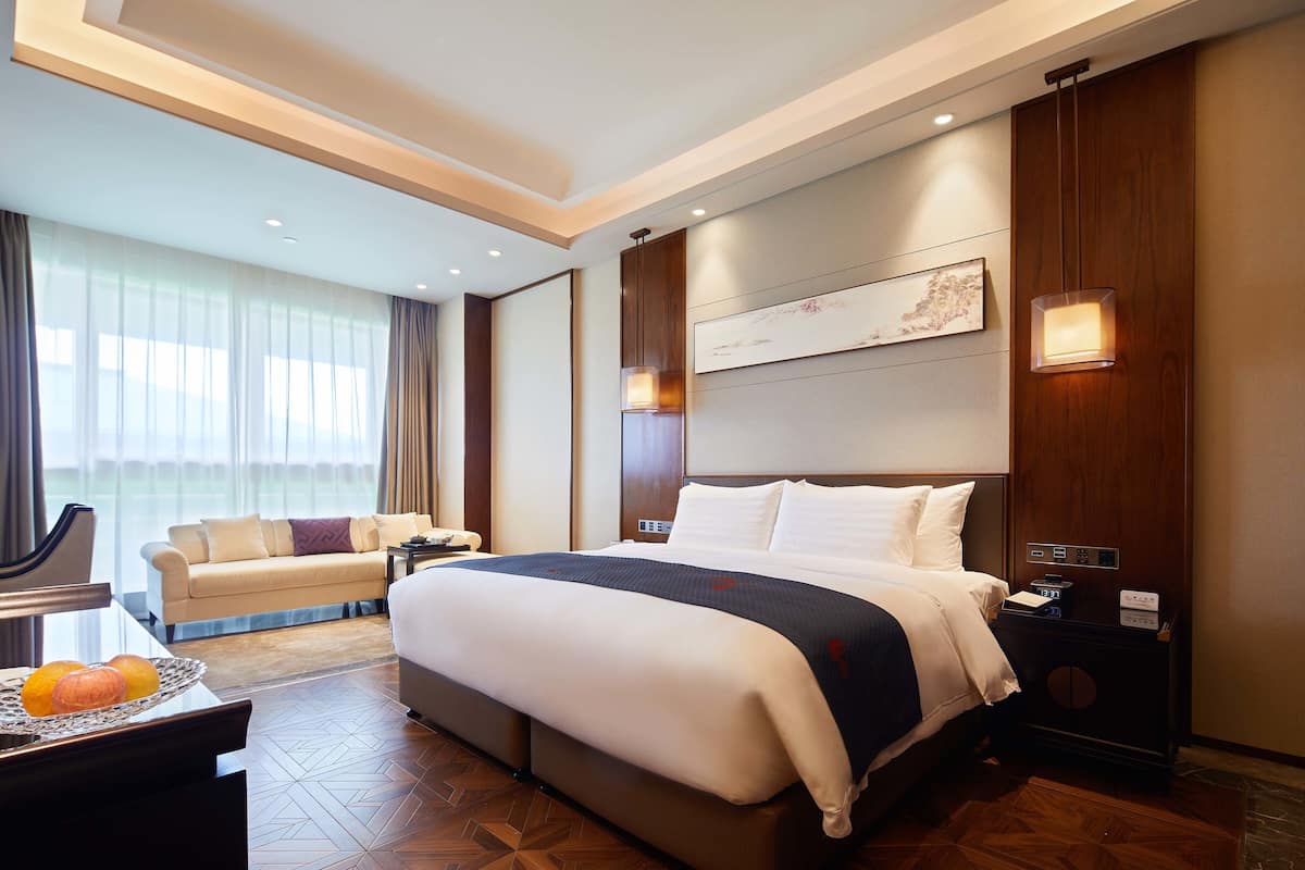 A hotel room with a large bed, a sofa, a desk with a tray of fruit, and a nightstand with a lamp.