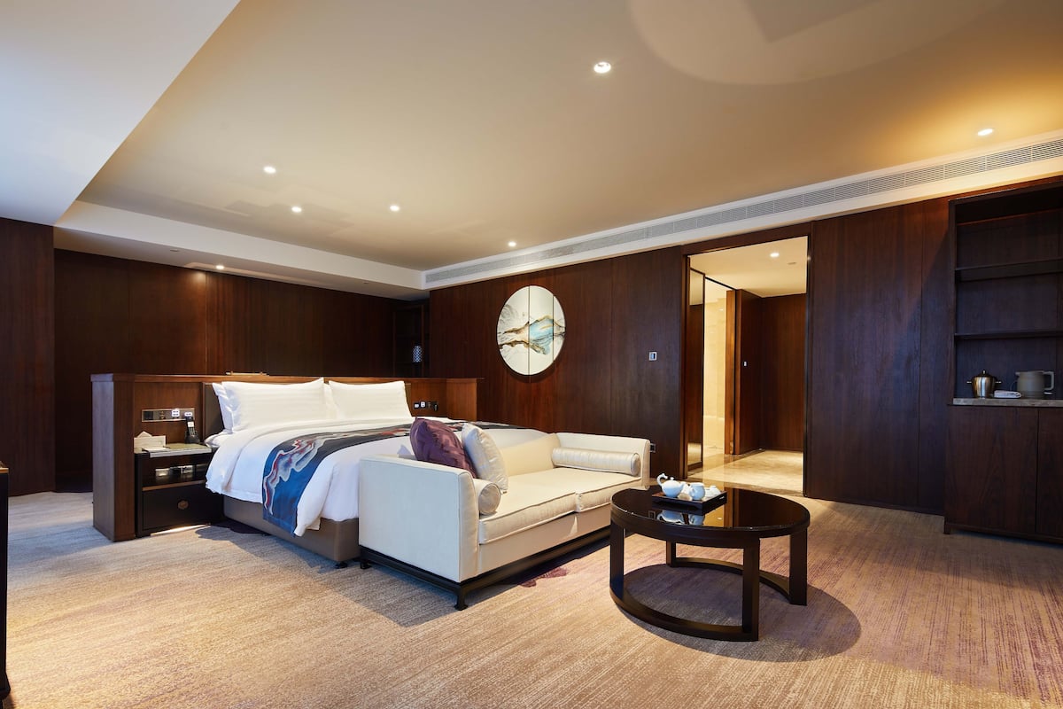 A modern hotel room with a bed, a sofa, a coffee table, and a large artwork on the wall.