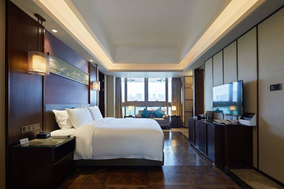 A hotel room with a large bed, bedside tables, a TV, and a view of the city through large windows.