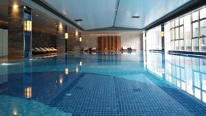 Una amplia piscina interior con suelo de baldosas, sillas de descanso e iluminación en la pared.