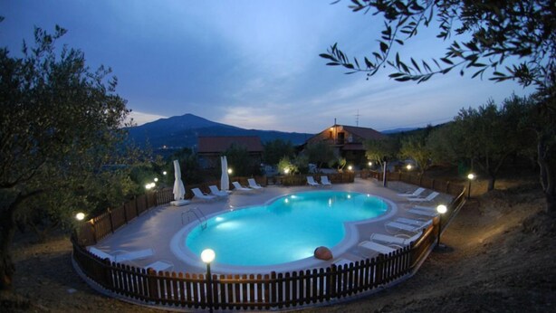 A circular swimming pool with a wooden deck and lounge chairs, surrounded by a fence and trees, with a mountain in the background. - Agriturismo Tempa del Niglio (Ascea)