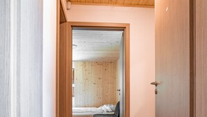 Un couloir étroit, doté d’un plancher en bois et d’un plafond percé d’une lucarne circulaire. Une porte située sur la droite donne accès à une pièce comprenant un lit et une petite table.