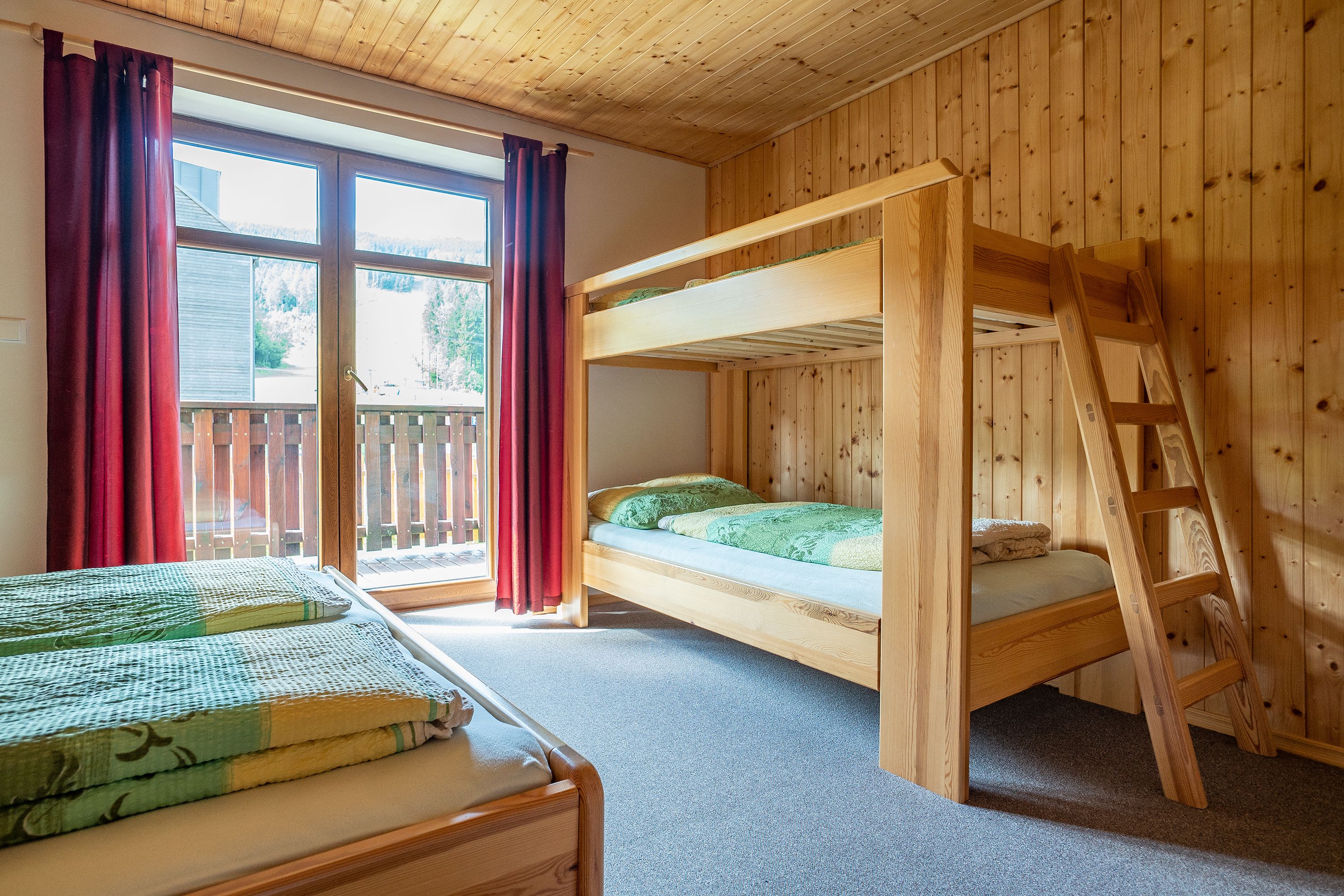 Habitación con literas, una escalera de madera, una ventana con cortinas rojas y vista a una terraza de madera.