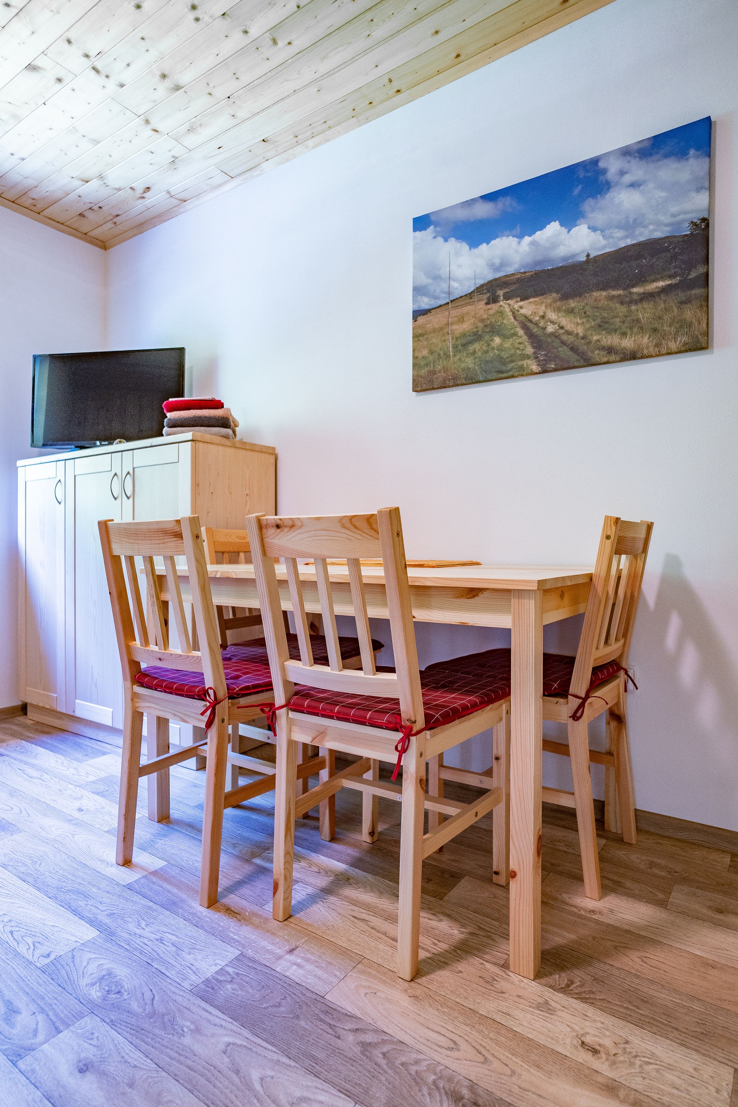 Un coin repas avec une table et des chaises en bois, un tableau au mur et un téléviseur posé sur un meuble.