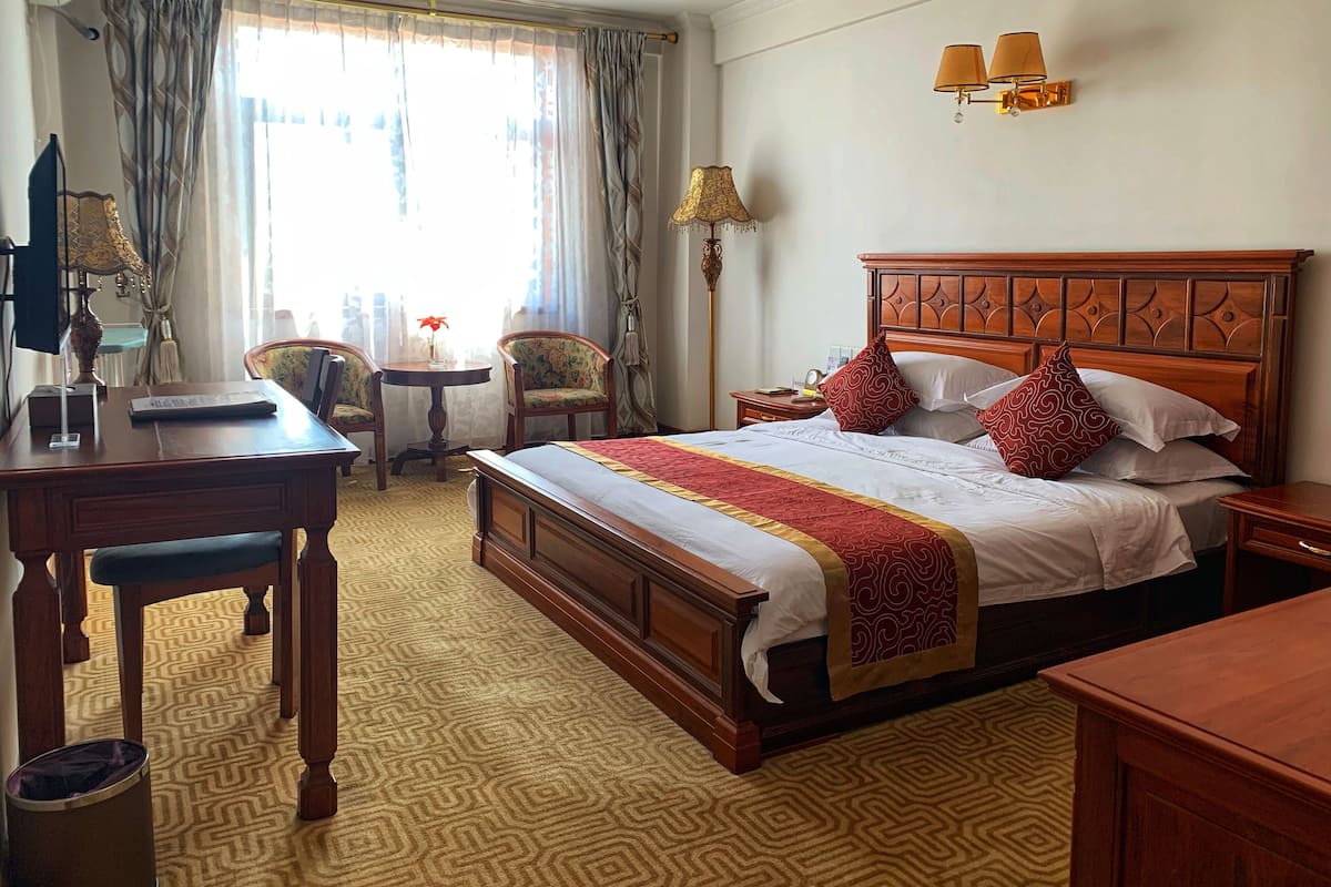 A hotel room with a large bed, a desk, two chairs, a small table with a lamp, and a window with curtains.