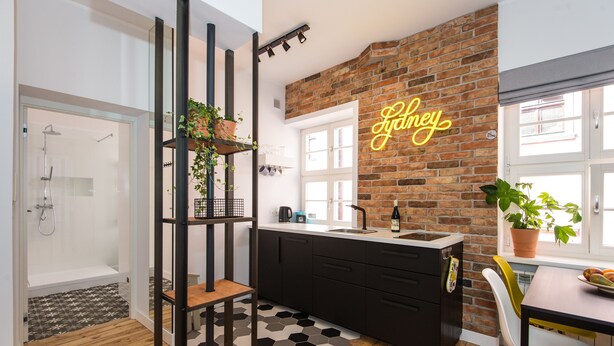 A modern kitchen with a brick wall, a neon sign, a sink, and a dining area with a plant on the table. - Bliss Apartments Sydney (Poznan)