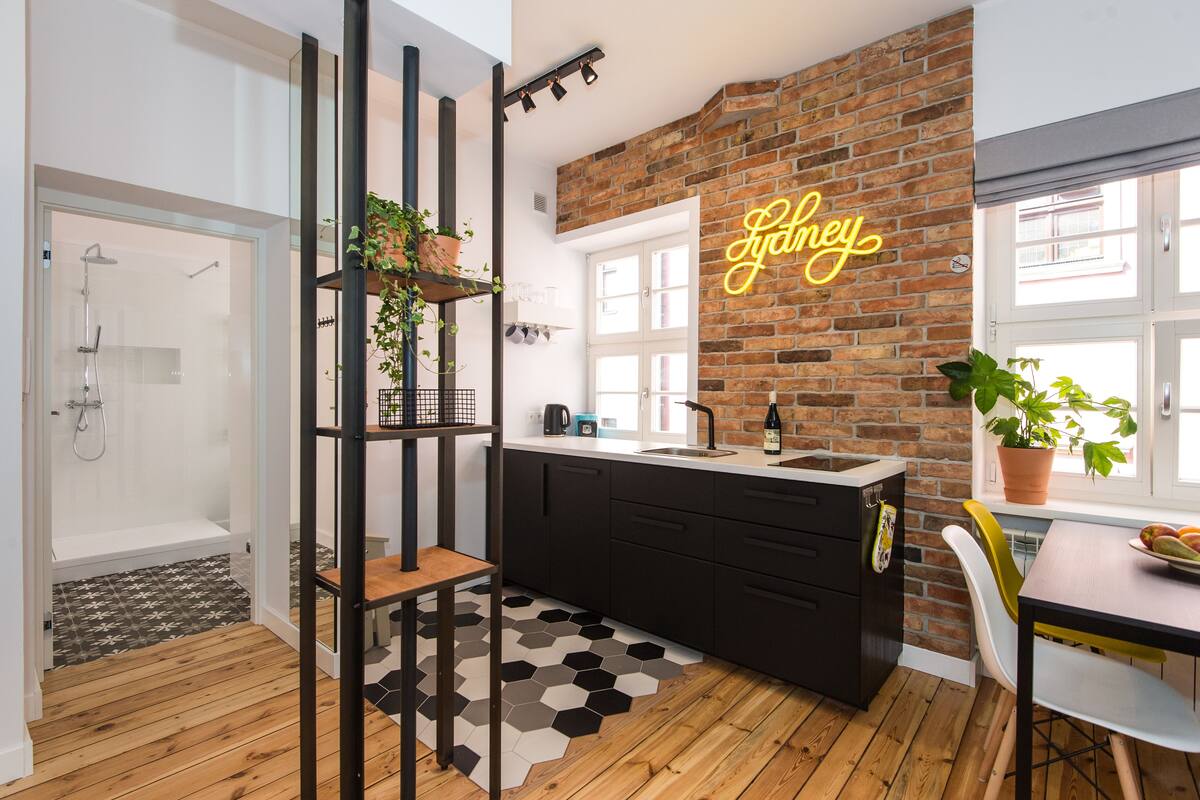 A modern kitchen with a brick wall, a neon sign, a sink, and a dining area with a plant on the table.