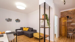 A modern living room with a grey sofa, yellow ottoman, wooden flooring, a black metal shelf with a plant, and a brick wall. - Bliss Apartments Sydney (Poznan)