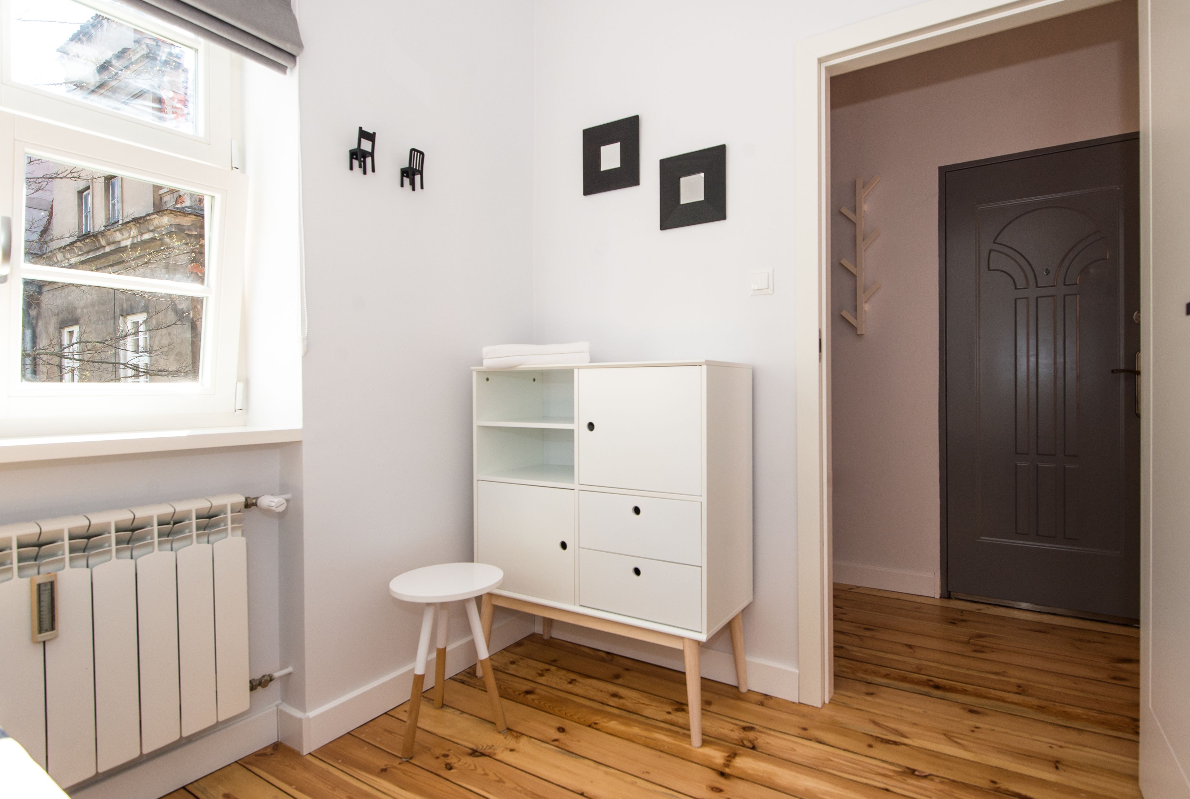 Una habitación con un radiador blanco, un armario blanco con cajones, un taburete pequeño y una puerta con un tirador decorativo.