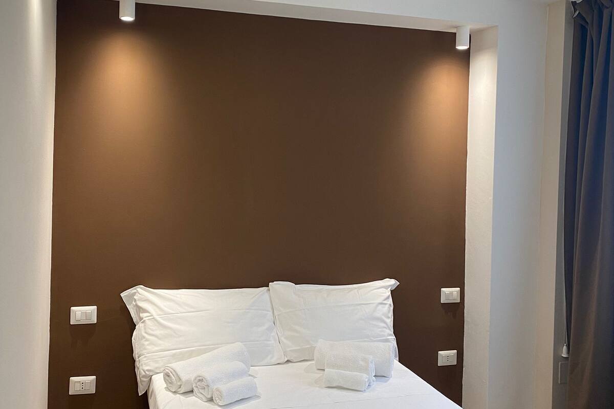 Habitación de hotel con cama, ropa de cama blanca, cabecera marrón y luz nocturna.