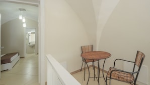 Un balcon avec une table et des chaises en osier, une balustrade blanche, et une vue sur une chambre à coucher avec un lit et une salle de bain en arrière-plan.