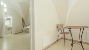 Une pièce avec un sol carrelé de carreaux blancs, un bureau blanc avec une chaise, une petite table ronde avec une chaise, et un mur blanc avec une porte.