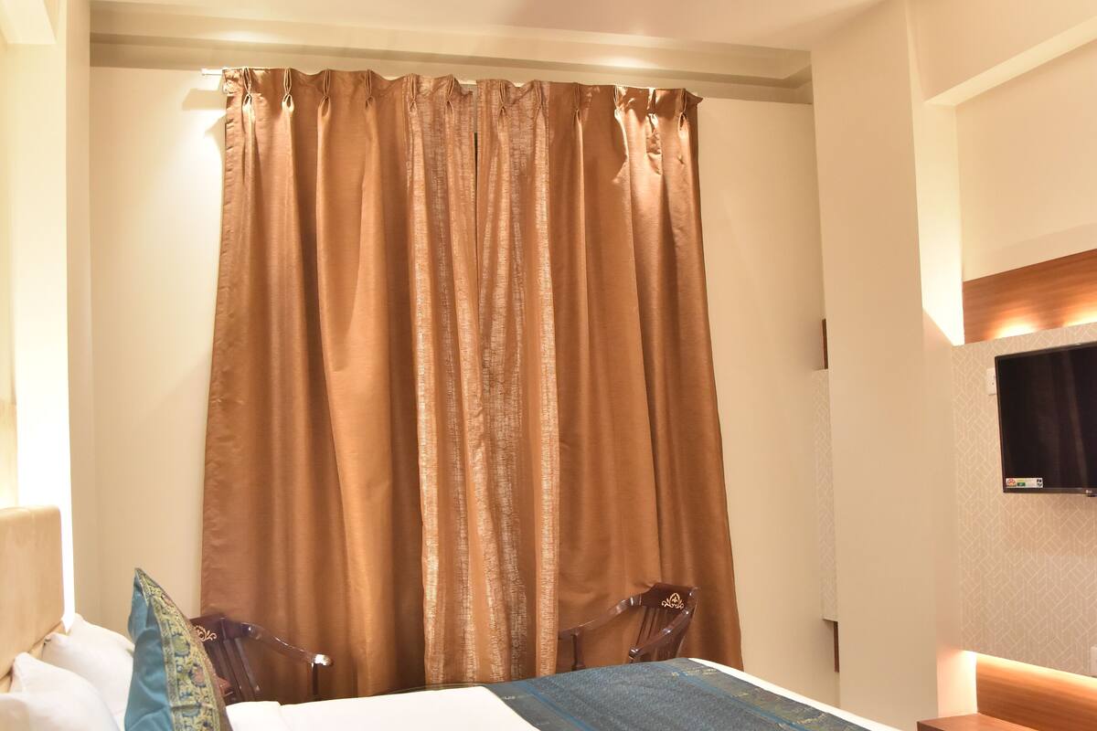 A hotel room with a bed, curtains, a ceiling fan, a mounted TV, and a wooden nightstand.