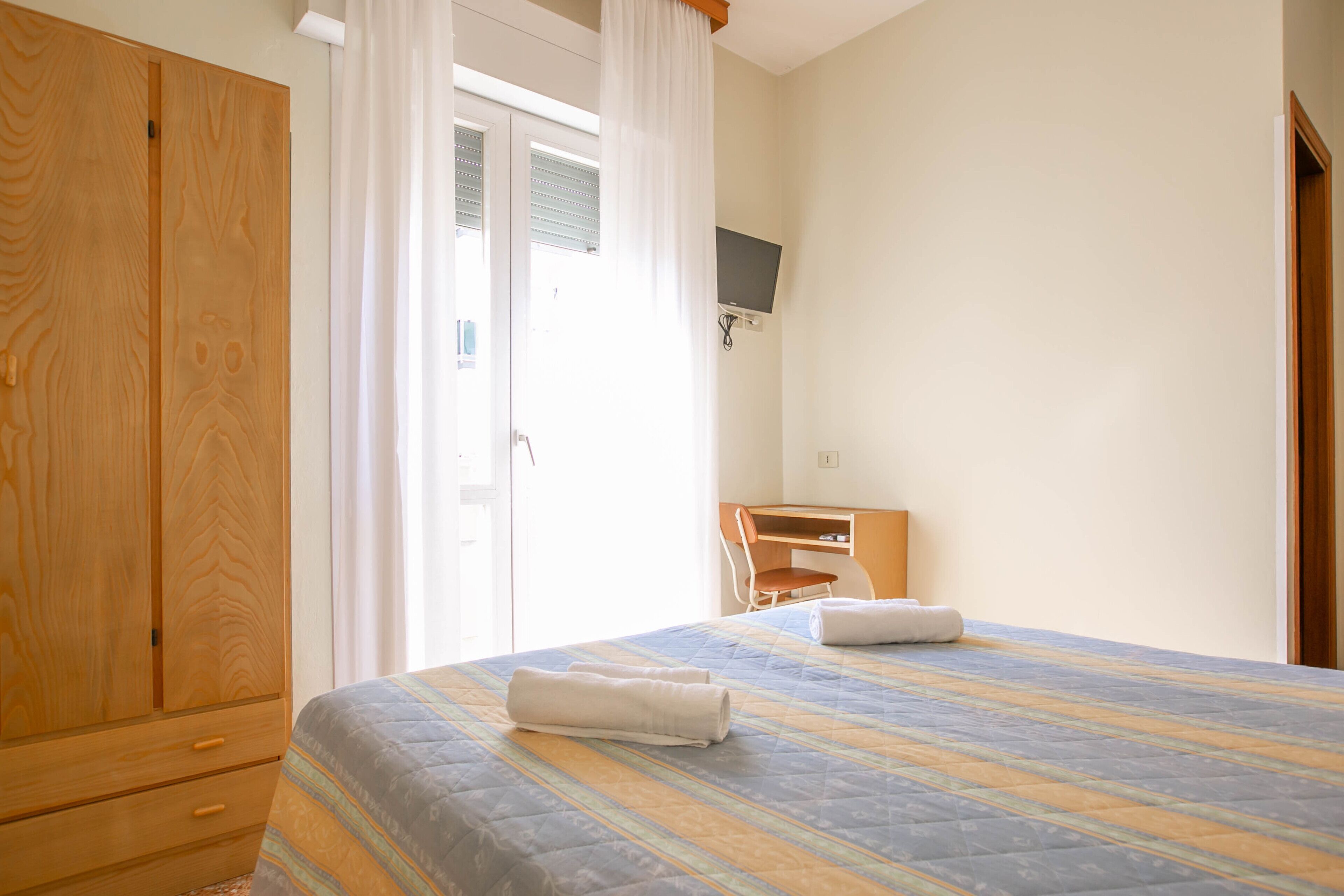 Een slaapkamer met een bed, een houten garderobe, een bureau met stoel, en een televisie aan de muur.