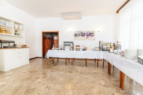 A spacious room with a long table set for a meal, a kitchen area with cabinets and shelves, and a door leading to another room. - Hotel Ischia (Rimini)