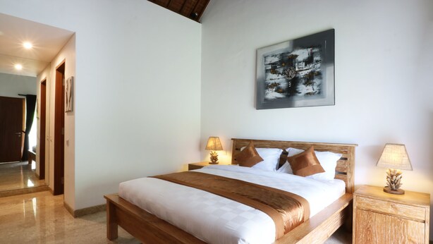 A hotel room with a large bed, wooden headboard, two bedside tables with lamps, a wall-mounted abstract painting, and a mirror in the corner. - Kubu Taulan (Kerobokan Kaja)