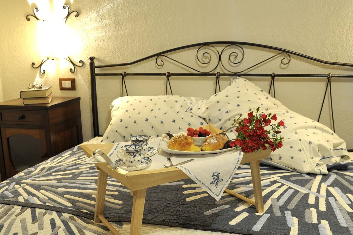 Una cama bien tendida con una colcha de estampado floral, una mesita de noche con una tetera y tazas, y una bandeja con pasteles y un jarrón de flores.