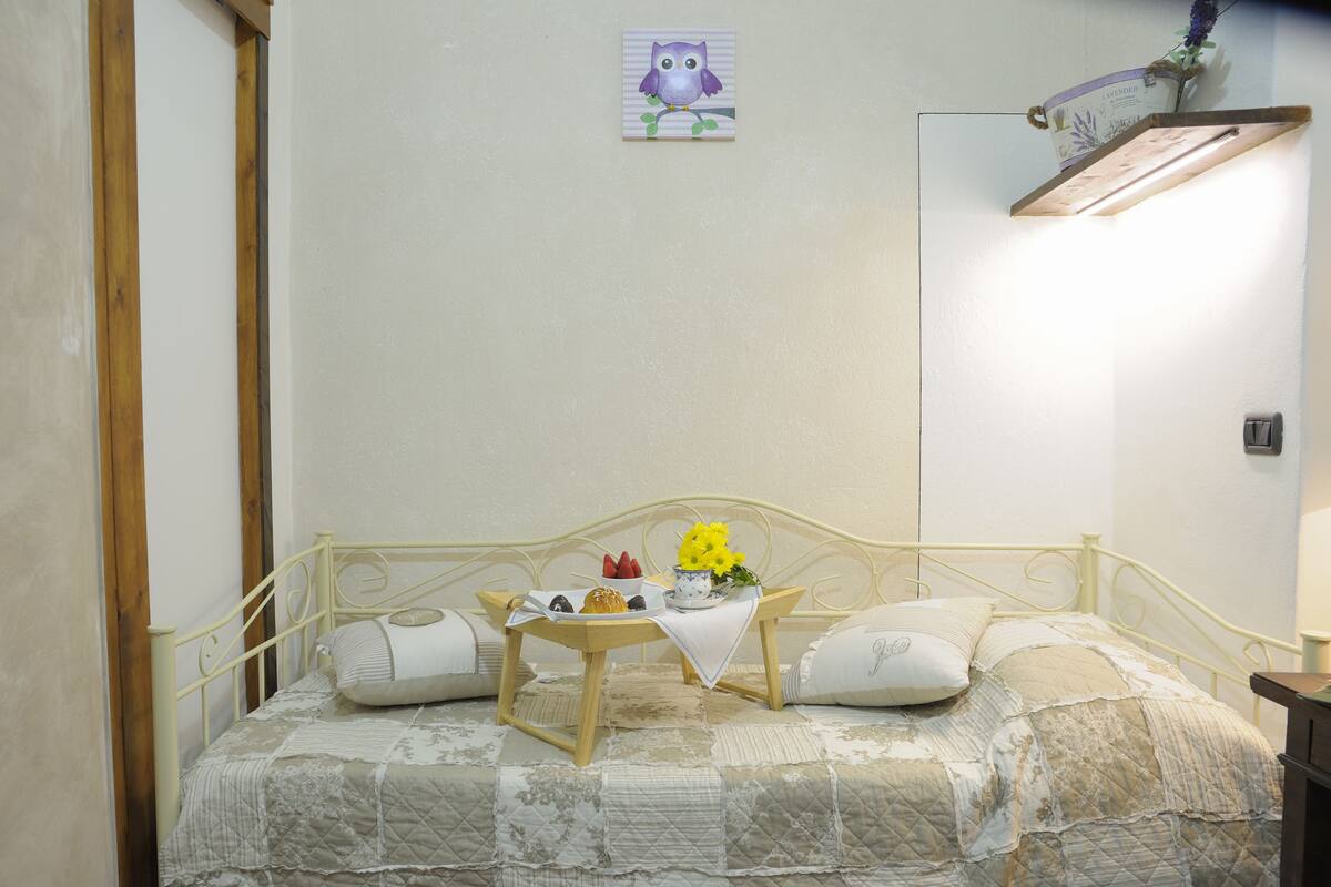 Una cama bien hecha con cubierta acolchada, una mesita de noche con una bandeja que sostiene un cuenco de flores, un plato con comida y una pequeña decoración de un búho en la pared.