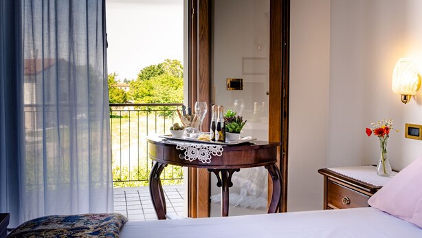 A bedroom with a bed, a bedside table with a vase of flowers, a nightstand, and a view of a balcony with a table set for a meal.