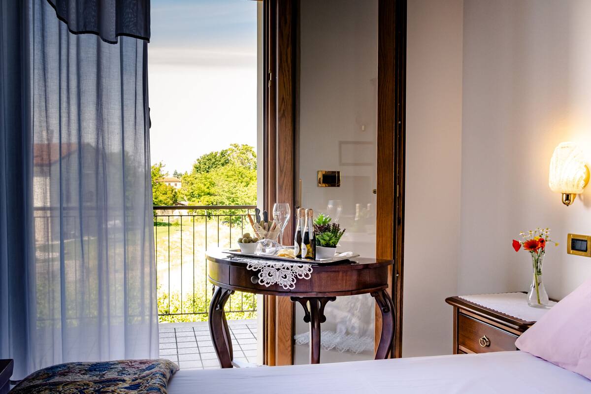 Un dormitorio con cama, una mesita de noche con un jarrón de flores, una mesita auxiliar y vistas a un balcón con mesa lista para comer.