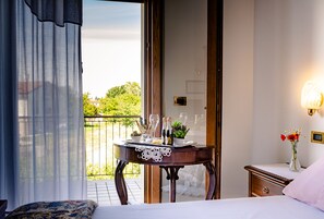Un dormitorio con cama, una mesita de noche con un jarrón de flores, una mesita auxiliar y vistas a un balcón con mesa lista para comer.