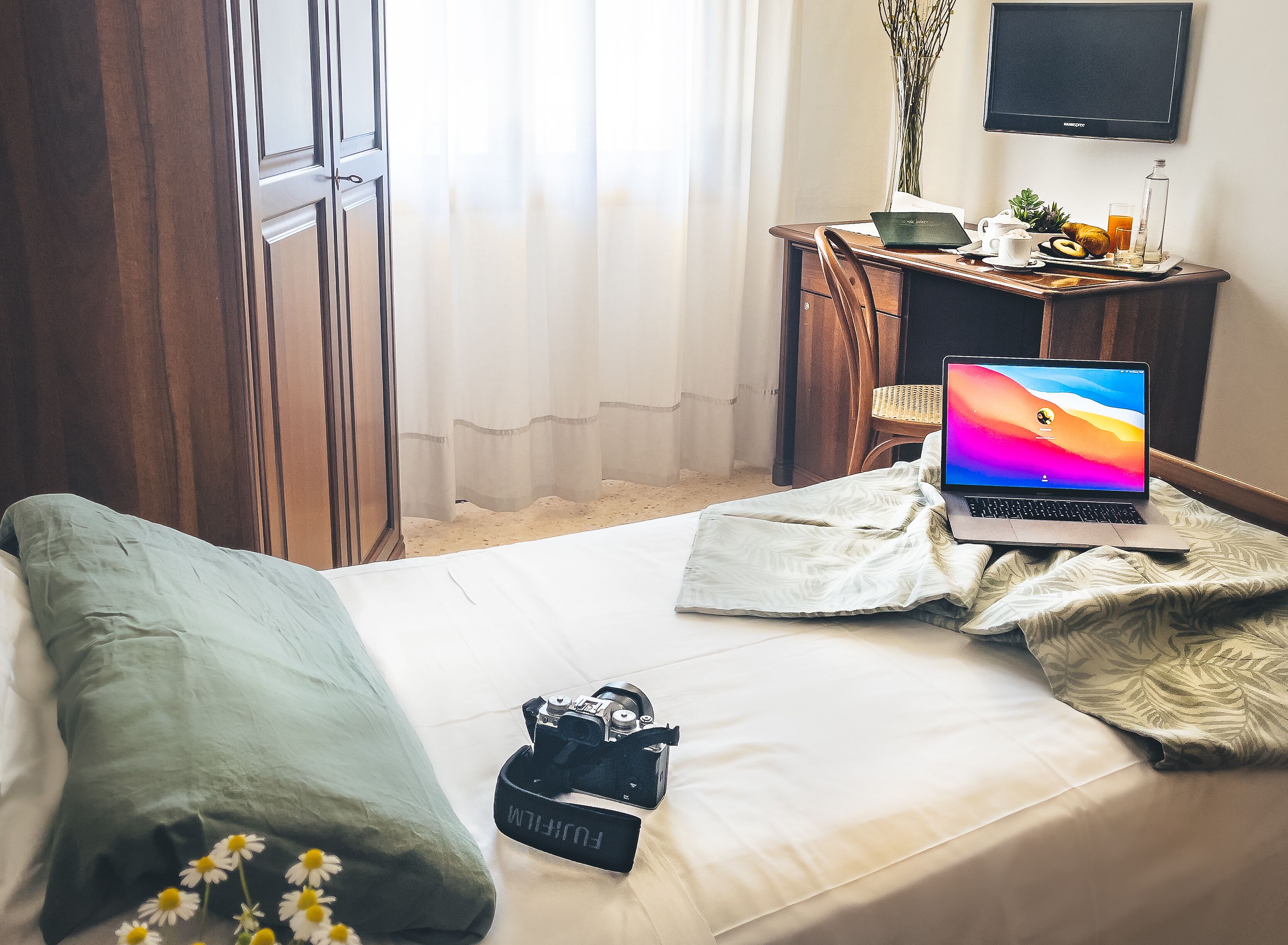 Une chambre d’hôtel avec un lit, un bureau avec un ordinateur portable, un appareil photo et un téléviseur fixé au mur.
