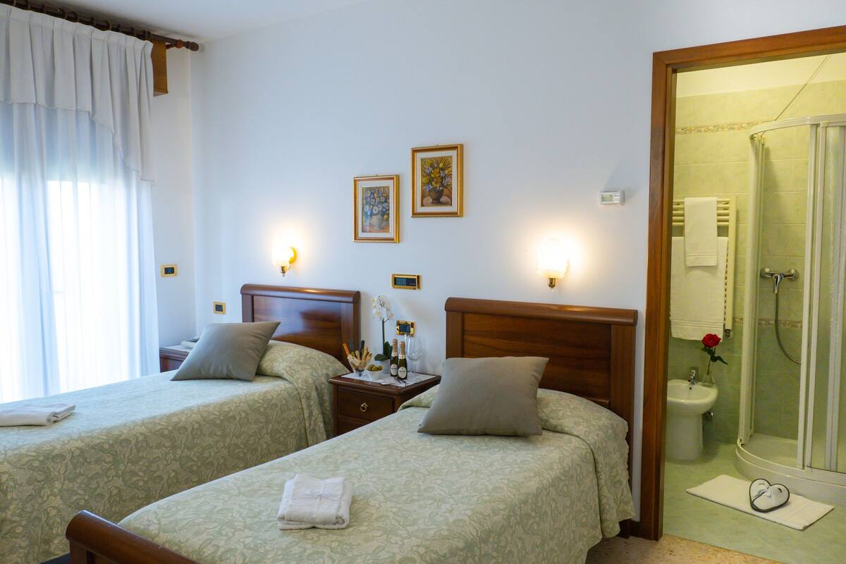 Habitación de hotel con dos camas, una mesita de noche con lámpara, baño con ducha y dos cuadros en la pared.