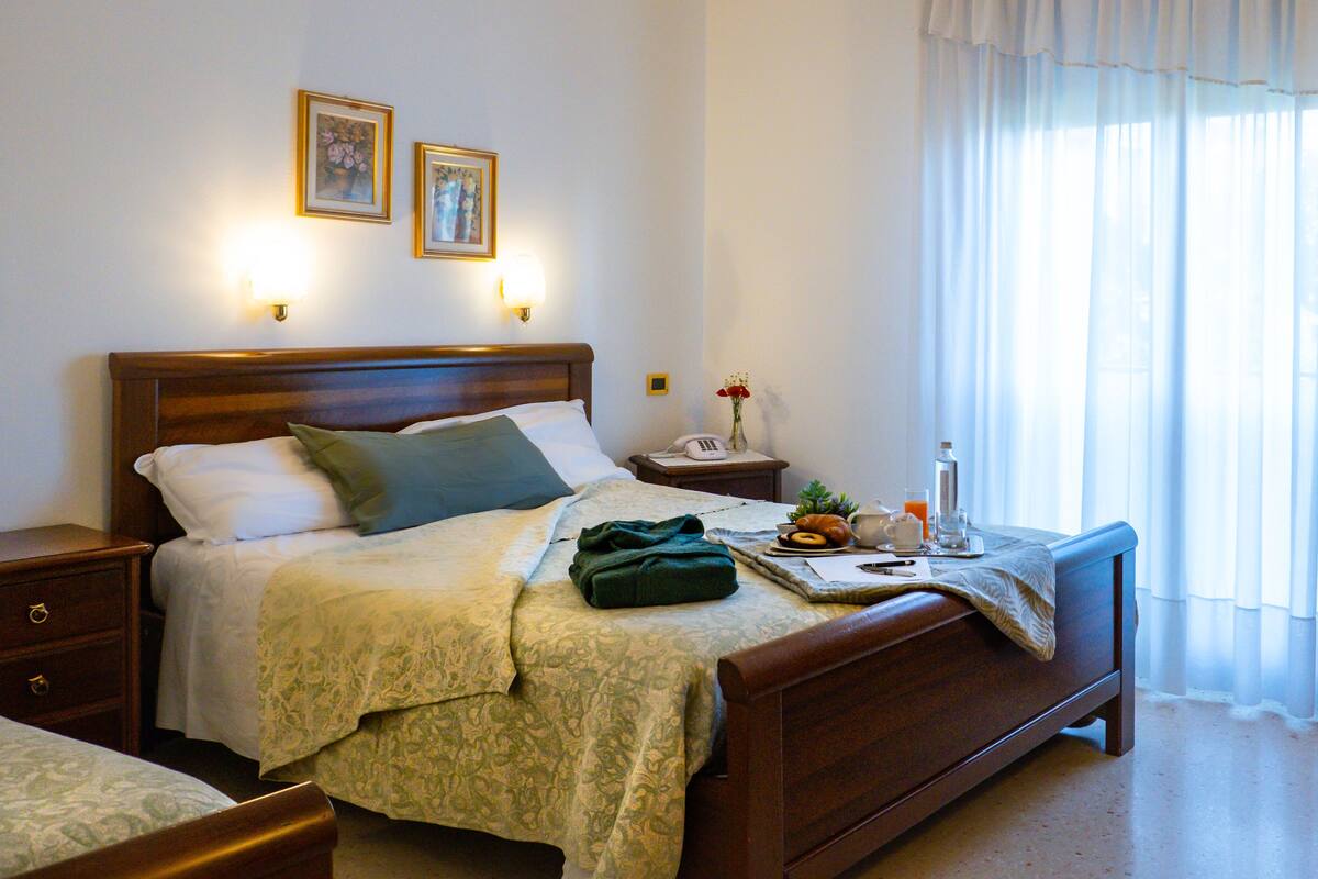 Una cama bien hecha con cabecera de madera, una mesita de noche con lámpara, un florero con flores y una bandeja de desayuno sobre la mesita.