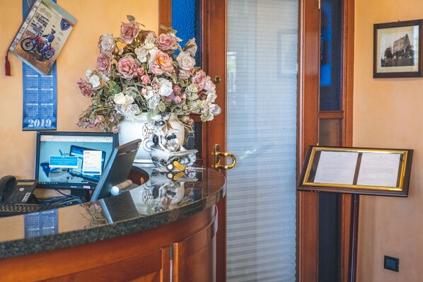 Un mostrador de recepción con una computadora, un florero, un calendario y una tablón de anuncios enmarcado.