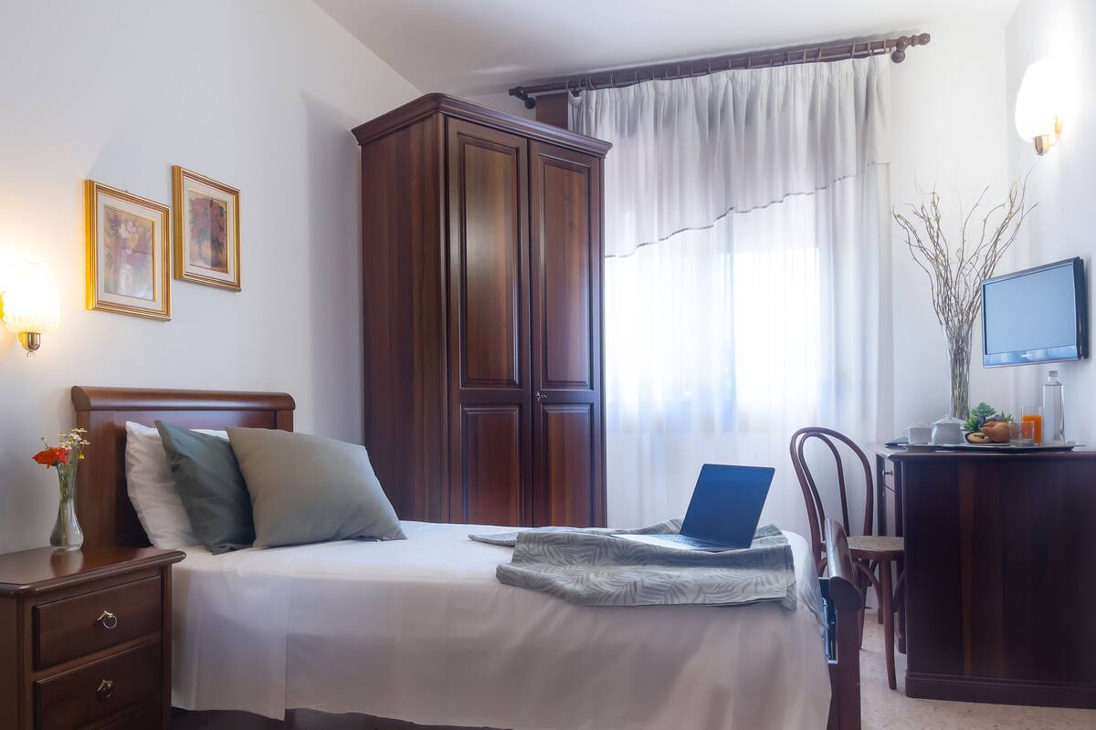 Un dormitorio ordenado con cama, mesitas de noche, armario, escritorio con una computadora portátil y televisor.