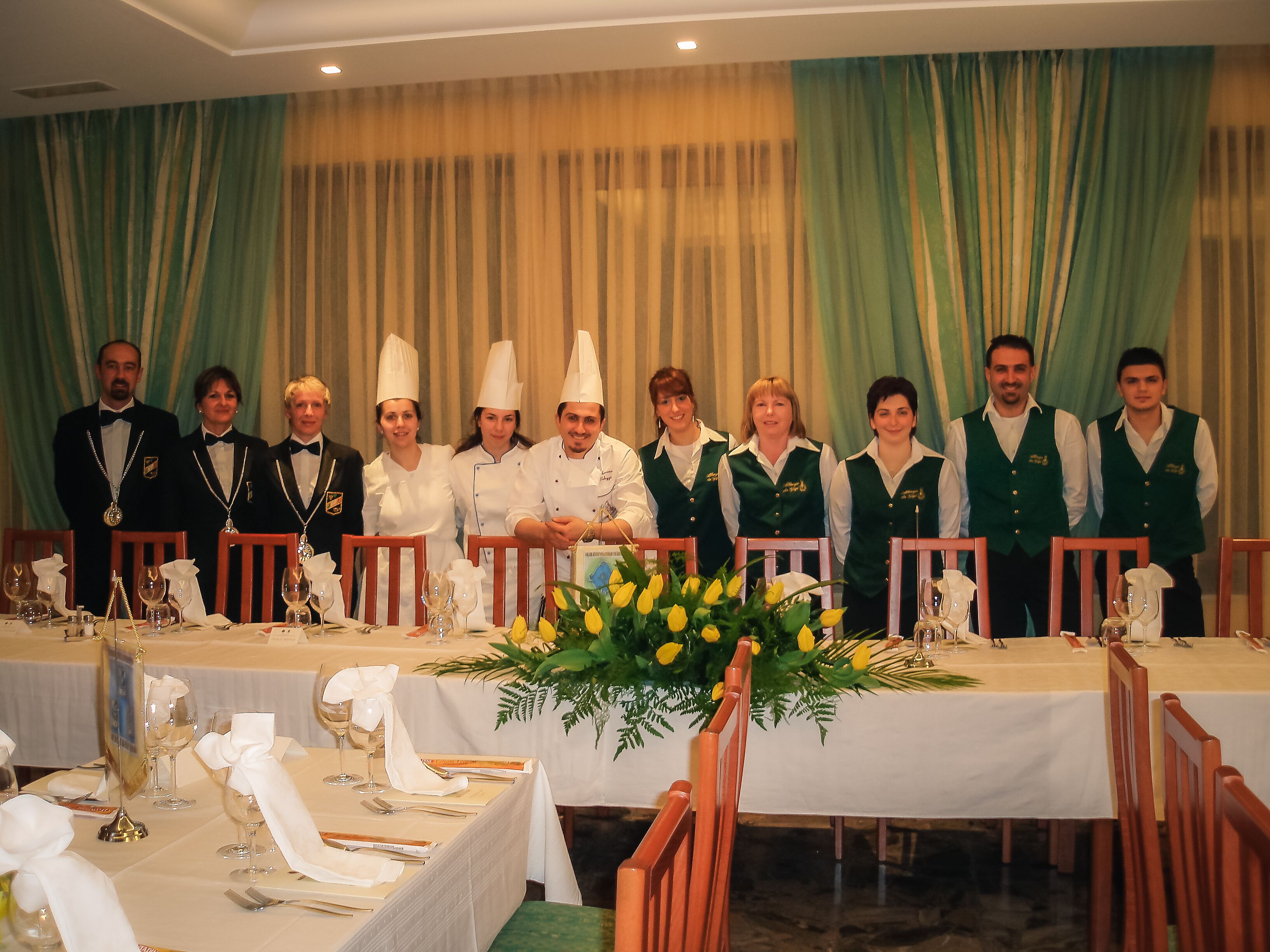Una elegante disposición para el comedor con mesas preparadas para una comida, con manteles blancos, cubertería ordenada y centros de mesa florales.