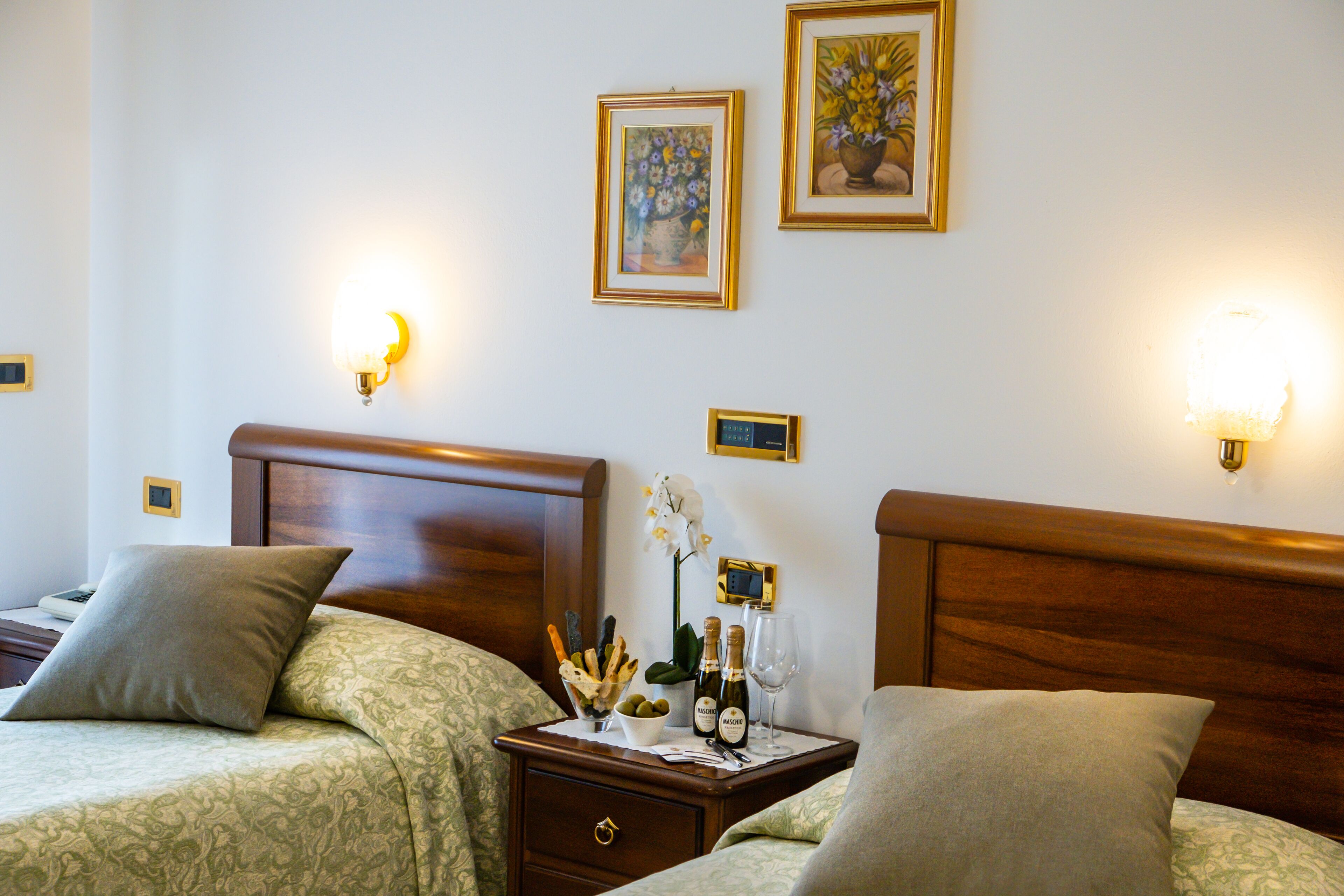 Ein Hotelzimmer mit zwei Betten, einem Nachttisch mit Blumenvase, Weinflaschen und gerahmten Bildern an der Wand.
