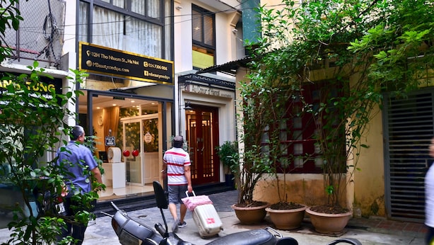 Vista de la calle con un edificio que muestra un letrero que dice 'Au Cœur d'Hanoi Apartments'. Hay plantas en macetas en la acera, una moto estacionada y una persona llevando una maleta.