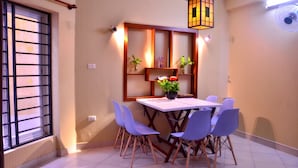 Un área de comedor con una mesa y sillas, una ventana con reja de metal, y un estante en la pared con plantas.
