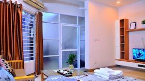 Habitación de hotel con cama, televisor, una pequeña mesa con un florero y control remoto, ventana con cortinas y estante con una planta.
