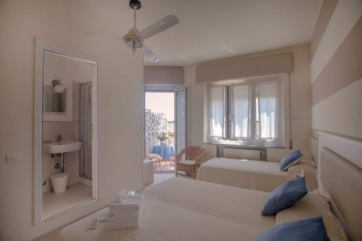 A bedroom with two beds, a window, a ceiling fan, and a bathroom visible through an open door.