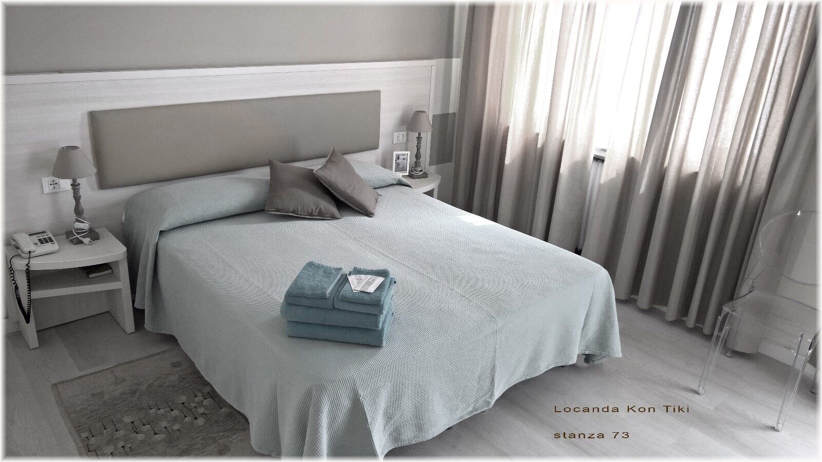 Una cama bien hecha con un cabecero gris, una mesita de noche con una lámpara y una toalla doblada sobre la cama.
