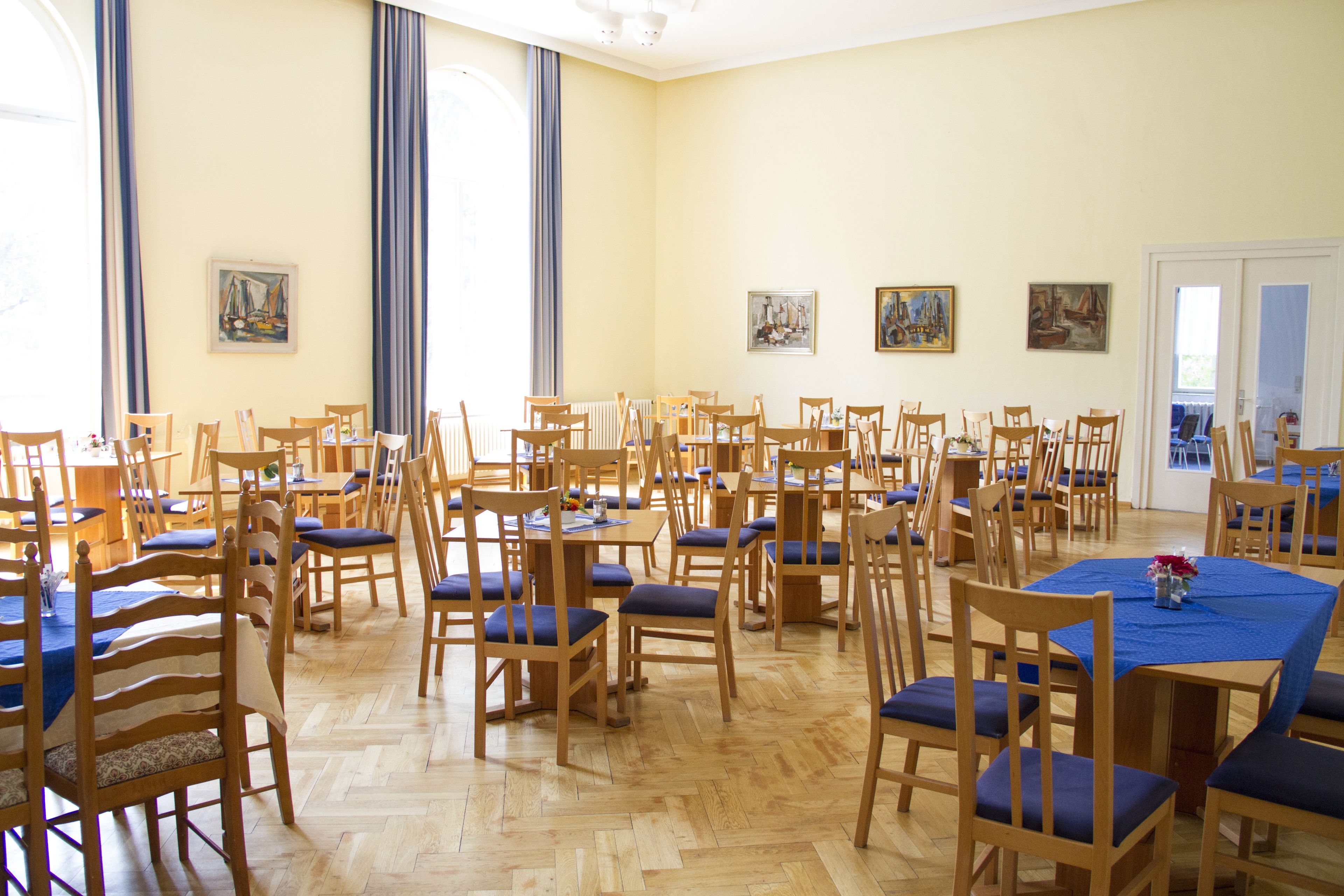 Ein geräumiger Speisesaal mit mehreren Holztischen und -stühlen, großen Fenstern mit blauen Vorhängen und verschiedenen Bildern an den Wänden.