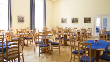 Ein geräumiger Speisesaal mit mehreren Holztischen und -stühlen, großen Fenstern mit blauen Vorhängen und verschiedenen Bildern an den Wänden.