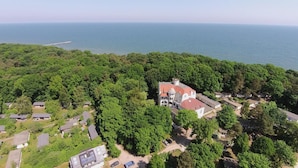 An aerial view of a coastal property with a large house surrounded by dense greenery, a pier extending into the sea, and a clear view of the ocean.