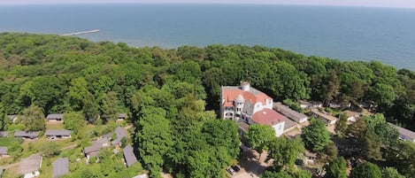 An aerial view of a coastal property with a large house surrounded by dense greenery, a pier extending into the sea, and a clear view of the ocean.