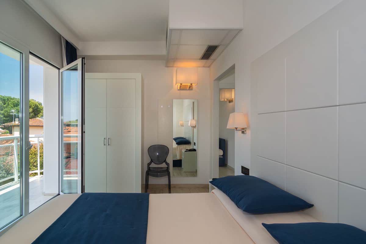 A modern hotel room with a large bed, a chair, a desk, and a balcony with a view of the city.