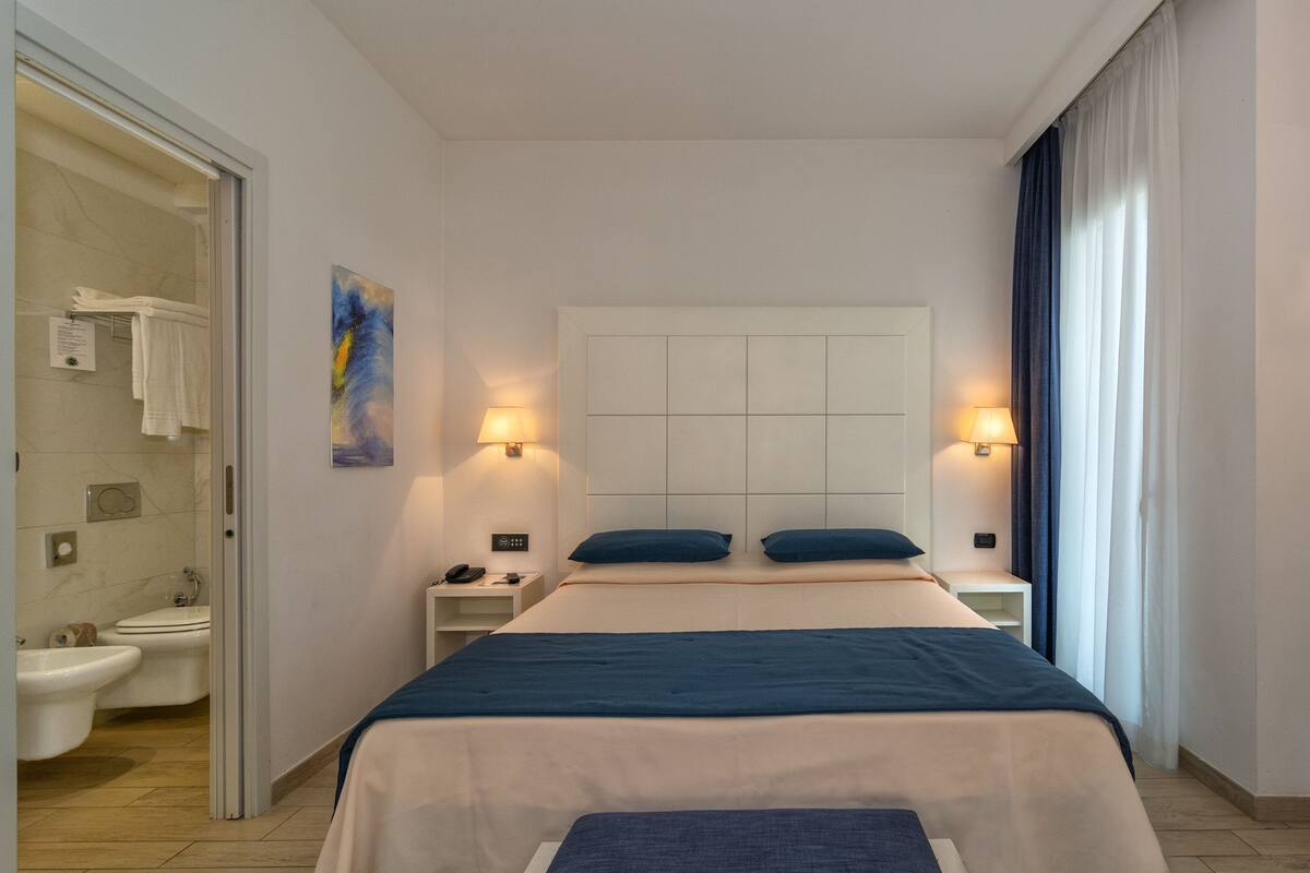A hotel room with a bed, bedside lamps, a nightstand, a bathroom with a toilet, bidet, and towel rack.
