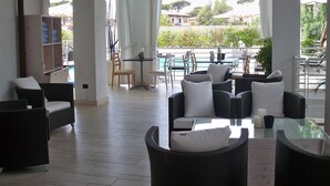 A spacious living area with wicker furniture, a glass table, and a view of a swimming pool and greenery. - Hotel Central Park (Pietrasanta)