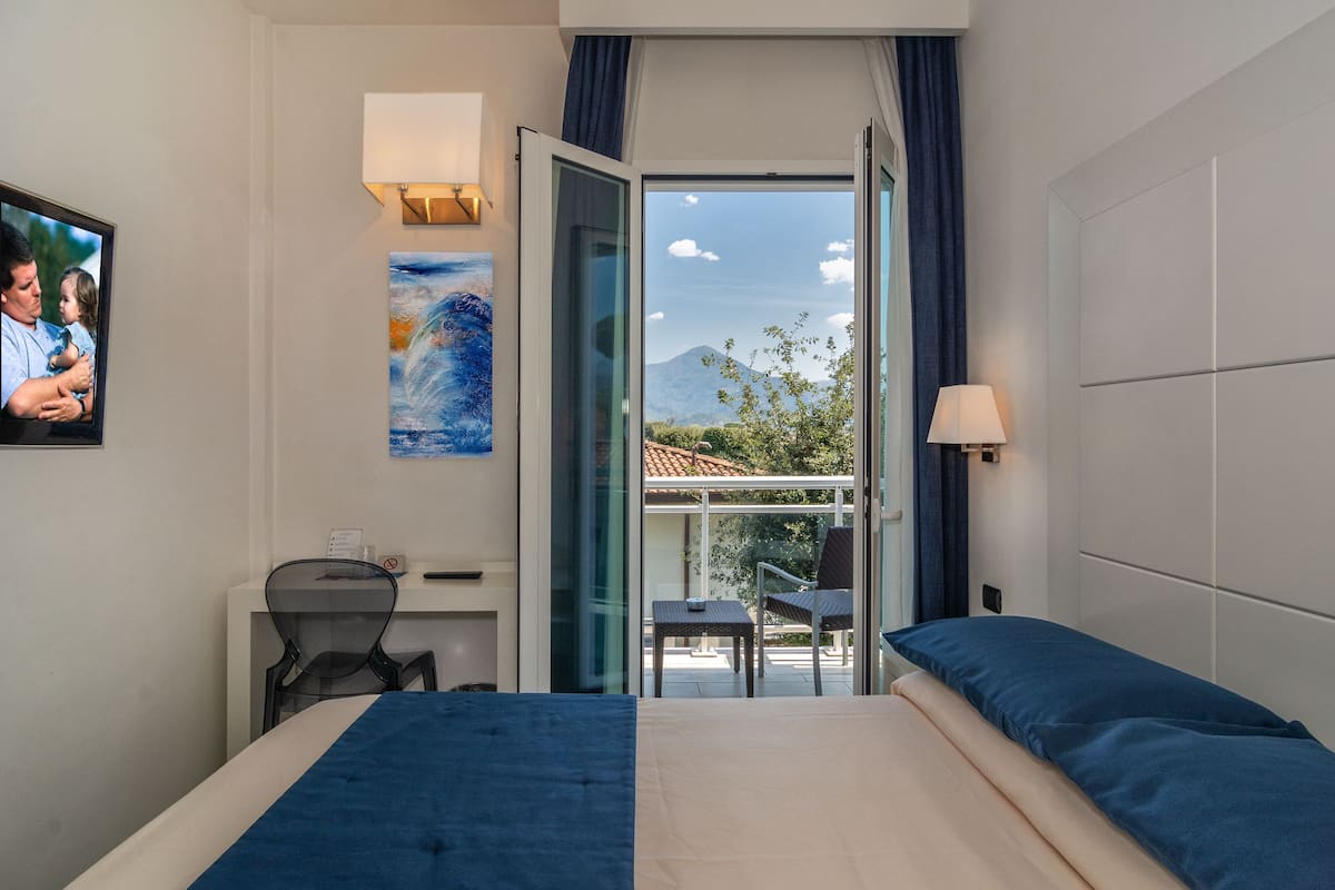 A hotel room with a bed, a desk, a chair, a TV, a painting, and a balcony with a view of a mountain.