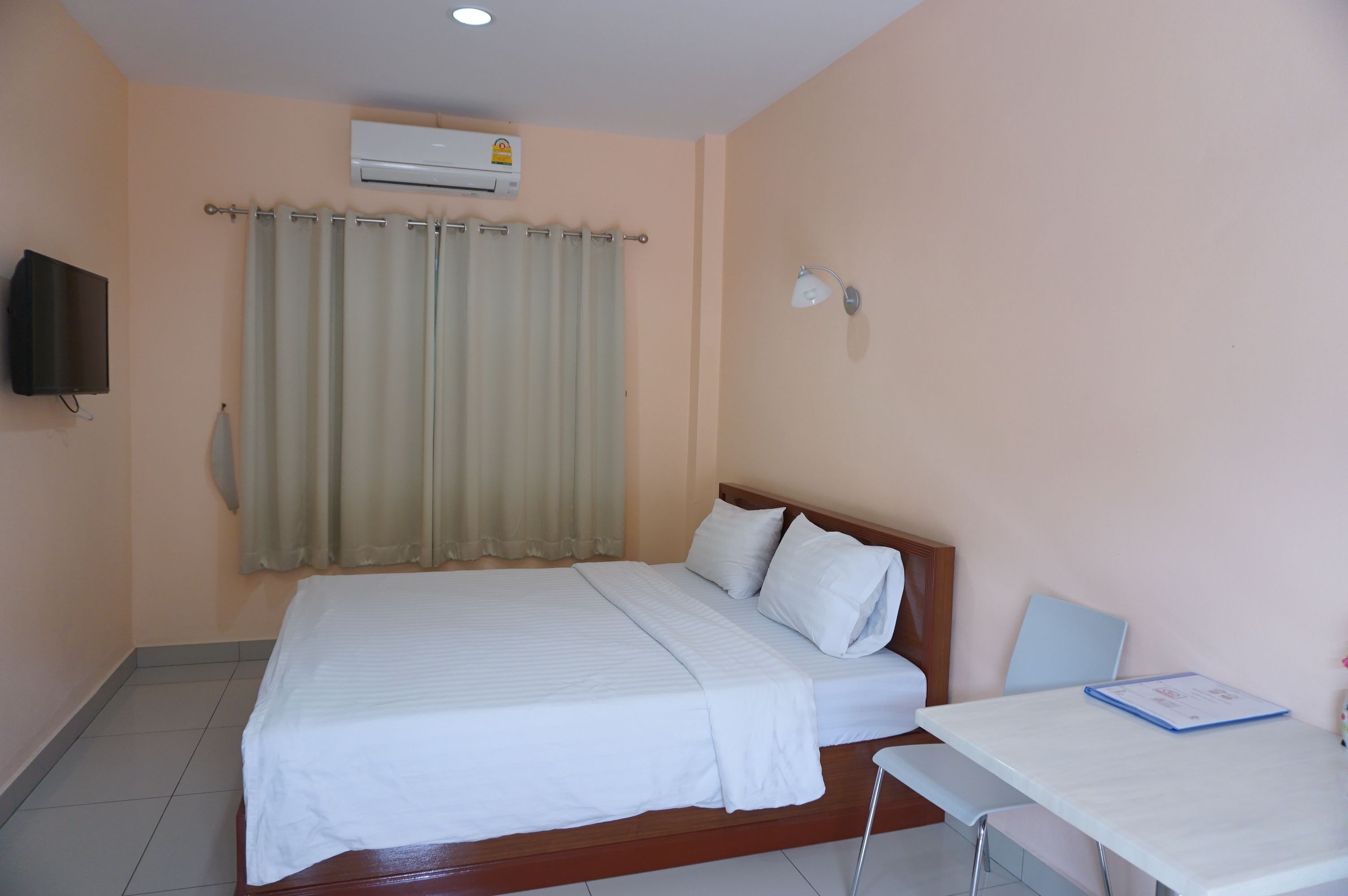A neatly made bed with white linens, a bedside table with a folder, a wall-mounted TV, and an air conditioning unit.