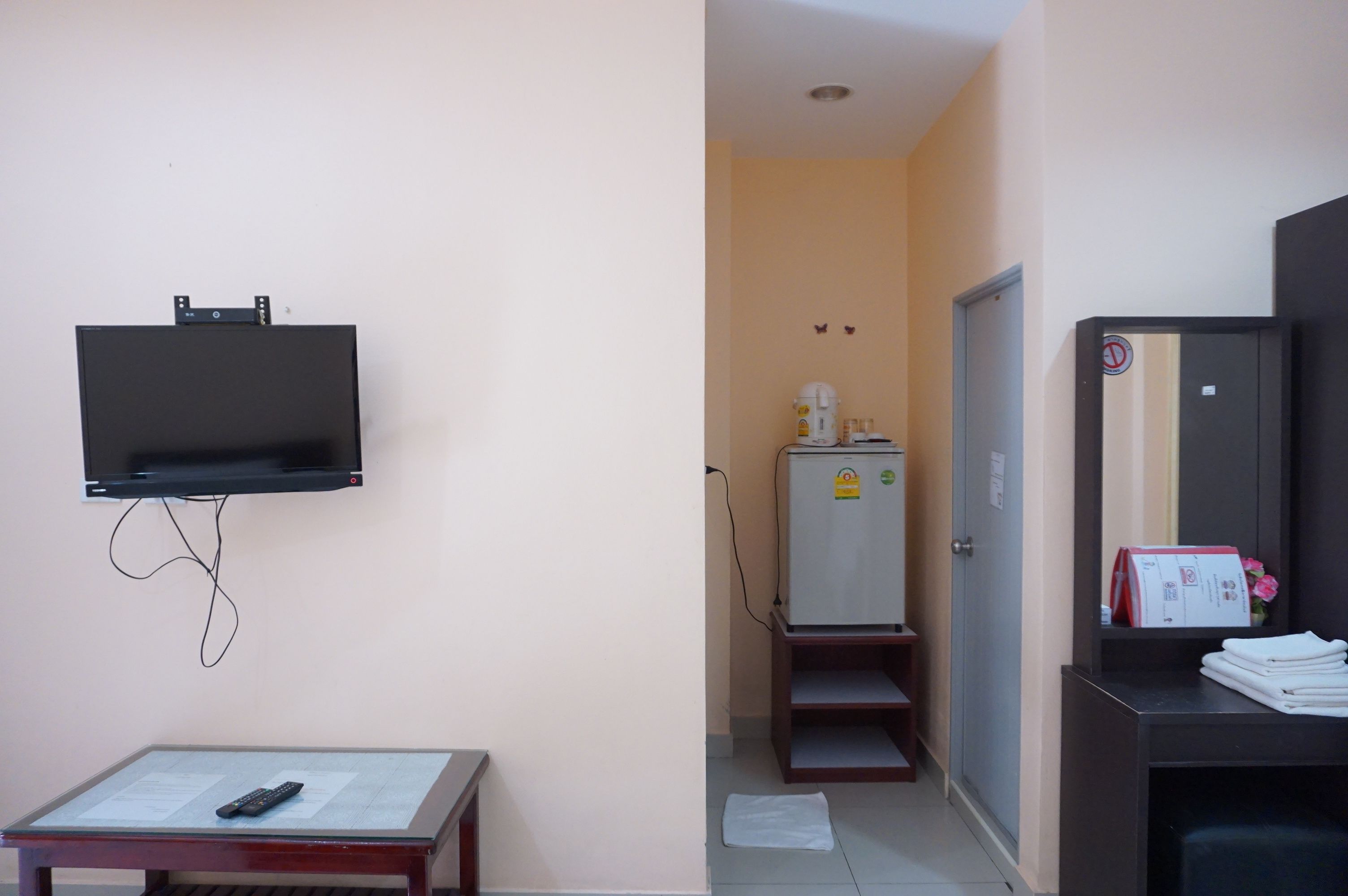 Habitación de hotel con televisión de pantalla plana en la pared, una mesita con un control remoto, un refrigerador y un escritorio con un espejo y unos papeles.